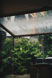 Terraza al aire libre en un día lluvioso, rodeada de arbustos verdes y vegetación. Gotas de lluvia caen sobre las plantas y el cerco, creando una atmósfera fresca y natural