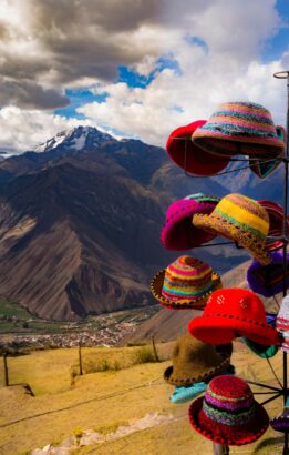Sombreros de colores colgando en primer plano, con las montañas de Cusco y un cielo con nubes de fondo